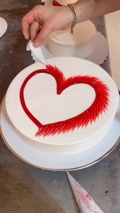 a person is decorating a heart shaped cake with red icing on a white plate