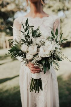 a woman in a wedding dress holding a white bouquet with greenery and flowers on it