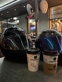 two cups of coffee sit next to each other on a table in front of a motorcycle helmet