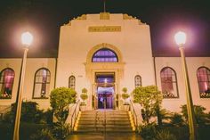 the front entrance to a building lit up by street lamps at night with potted plants on either side