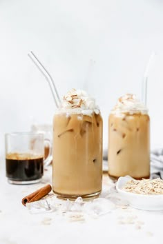 two glasses filled with iced coffee on top of a white table next to cinnamon sticks