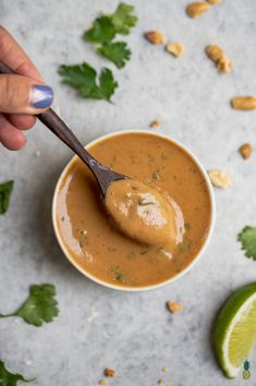 a hand holding a spoon full of peanut sauce with limes and peanuts around it