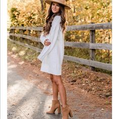 a woman wearing a white sweater dress and brown hat standing in front of a fence