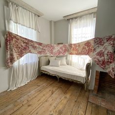 a bed sitting on top of a wooden floor next to two windows with drapes