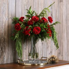 a glass vase with red roses and greenery on a wooden table next to a fan