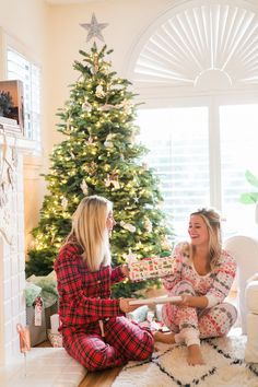 two women sitting on the floor in front of a christmas tree, one holding a present