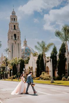 a bride and groom walking across the street in front of a church with palm trees