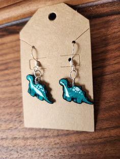 a pair of blue dinosaur earrings sitting on top of a wooden table next to a tag