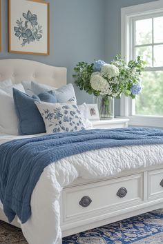 a white bed with blue and white pillows in a bedroom next to a vase filled with flowers