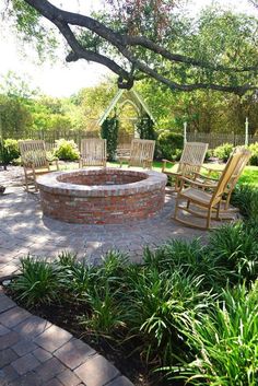 a brick fire pit surrounded by lawn chairs and greenery in the back yard area