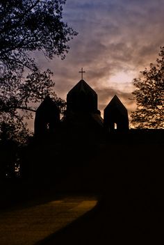 the silhouette of two church towers against a cloudy sky at sunset with trees in foreground