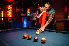 a man and woman leaning over a pool table with balls in front of them at night