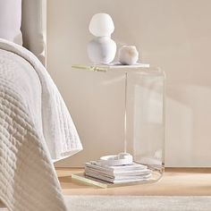 a bed with white sheets and pillows next to a glass shelf on the side table