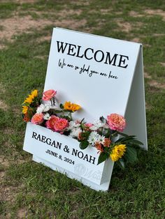 a welcome sign sitting in the grass with flowers on it's side and an inscription that reads, welcome we are helping me here