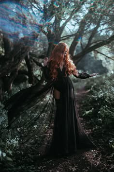 a woman with long red hair in a black dress is walking through the woods and has her arms outstretched