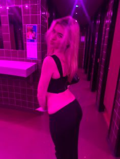 a woman standing in a room with pink lights on the walls and wearing tight black pants