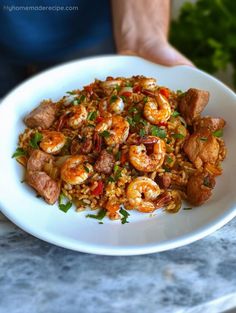 a white bowl filled with shrimp and rice on top of a marble counter next to a person's hand