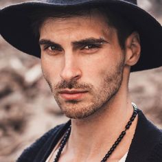 a young man wearing a black hat and beaded necklace looks at the camera while standing in front of some rocks