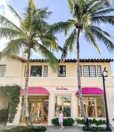 a woman standing in front of a pink and white building with palm trees on the sidewalk