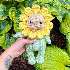 a hand holding a crocheted sunflower doll in front of some green leaves