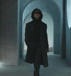 a man in a black coat and sunglasses walking down a hallway with an archway behind him