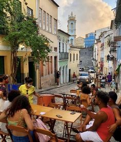 several people sitting at tables in an alleyway with buildings and cars on the street