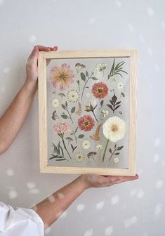 a person holding up a framed painting with flowers on it's side and white dots in the background