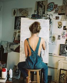 a woman sitting on top of a stool in front of a easel filled with art