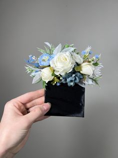 a hand holding a small black box with white and blue flowers in it's center