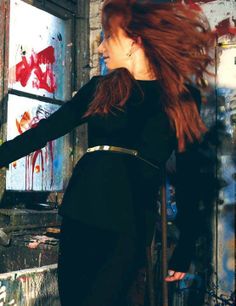 a woman with long red hair standing in front of a graffiti covered wall wearing a black dress