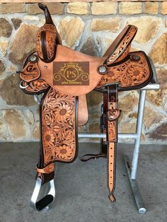 a brown and black horse saddle sitting on top of a metal stand next to a stone wall