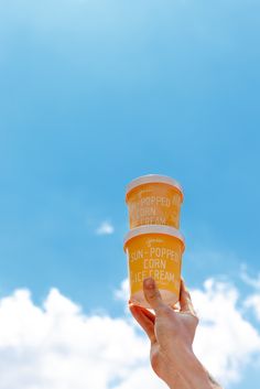 someone holding up two cups of ice cream in front of a blue sky with clouds