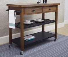 a wooden table with two drawers and a shelf on wheels holding coffee cups, teacups and books