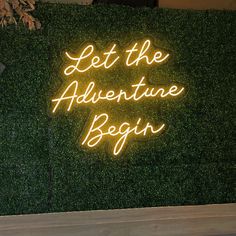 a neon sign that says let the adventure begin on top of a green hedge wall