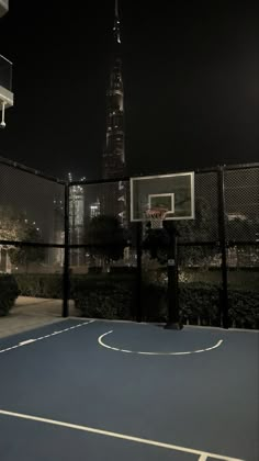 an empty basketball court in front of a tall building at night with the lights on