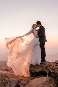 a bride and groom standing on top of a mountain at sunset with their arms around each other