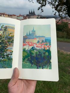 an open book with watercolors on it in the grass near trees and buildings