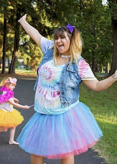 a woman in a tie dye skirt and jean jacket is holding her arms up as she walks down the road