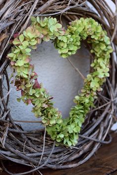 a heart shaped wreath made out of twigs and green leaves on a wooden table with a white vase in the background