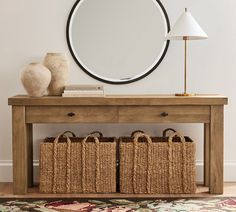 a wooden table with two baskets under a round mirror on the wall next to a lamp