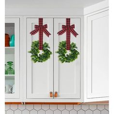 two wreaths are hanging on the front of white cabinets with red bows and green ivy