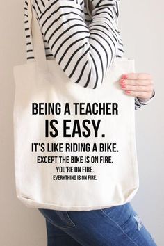 a woman holding a tote bag that says being a teacher is easy it's like riding a bike except the bike is on fire you're on fire everything