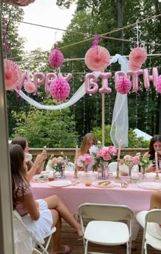 a group of women sitting around a table with pink decorations on it and the words happy birthday hanging above them