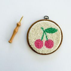 an embroidered cherry ornament next to a sewing needle and wooden spool on a white surface