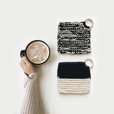 a person holding a cup of coffee next to two knitted coasters on a white surface