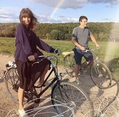 two people riding bikes on a dirt road with a rainbow in the sky behind them