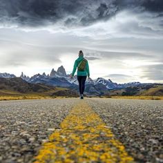 a woman is walking down the road with mountains in the backgrouds behind her