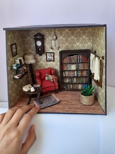 a hand is holding a dollhouse with a red chair and bookshelf in it