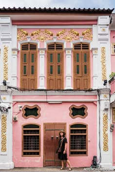 a woman standing in front of a pink and white building with wooden shutters on the windows