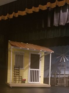 a small yellow house sitting on top of a stage next to a wooden chair and table